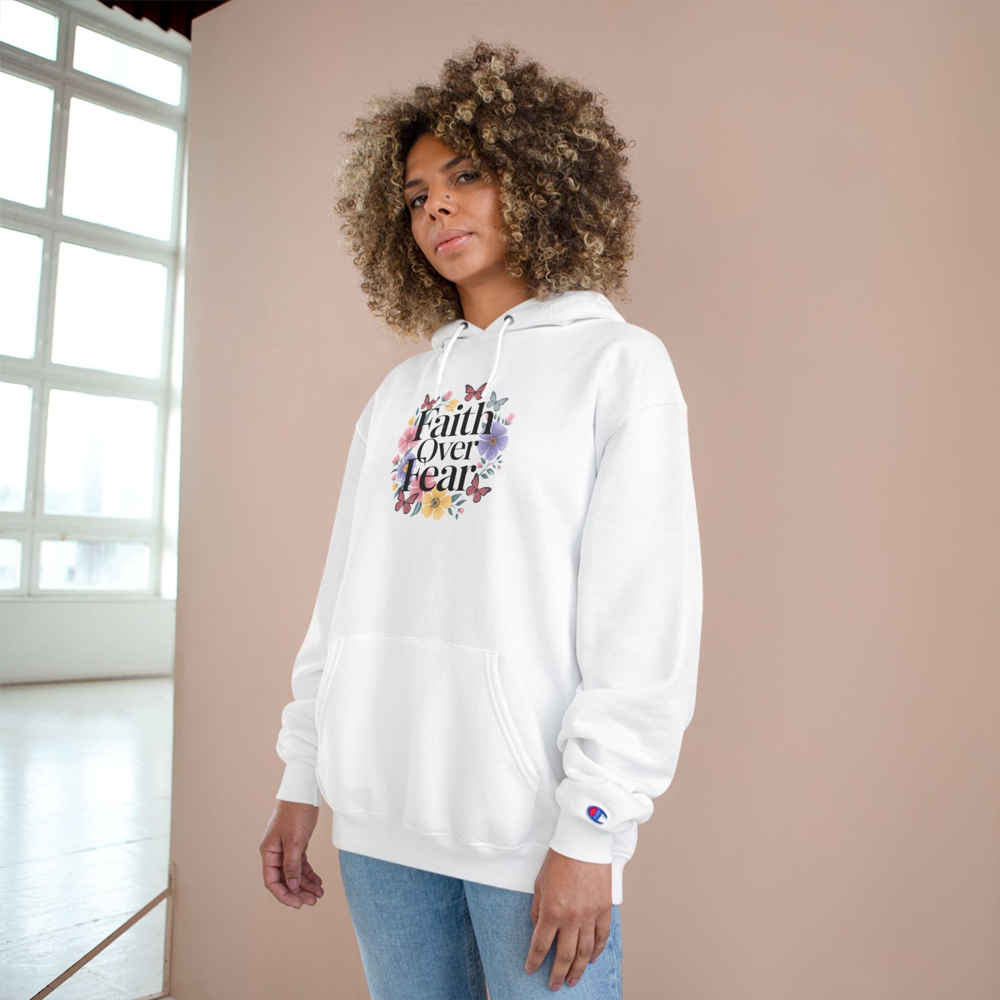 Divine Faith Armor™ “Faith Over Fear” Champion Hoodie with bold faith design and uplifting message Woman wearing a white hoodie with 'Faith Over Fear' text and floral design against a beige wall.